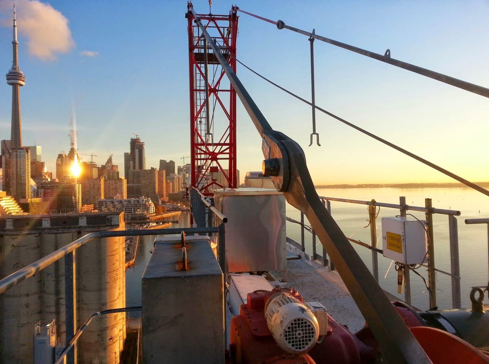 Hoist motor on a red tower crane above the Toronto waterfront at sunrise, the CN Tower in the distance